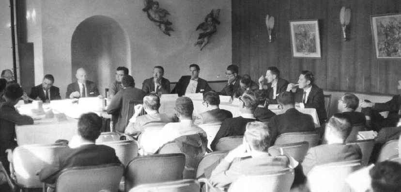 A black-and-white photograph of about twenty men seated around a long U-shaped table in a conference room. Attendees in suits sit side by side at the far table, while those in the foreground face them with their backs to the camera. Framed pictures and decorations hang on the walls.